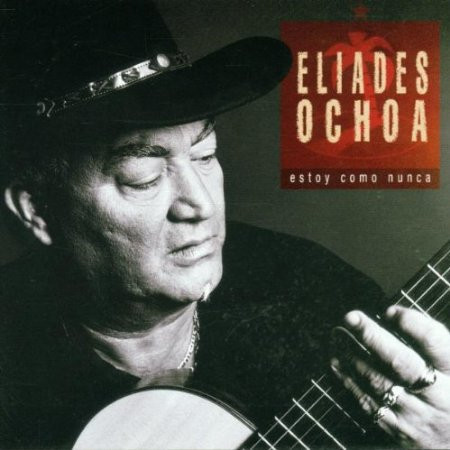 The face of Eliades Ochoa playing the guitar, his lowered eyes looking at his left hand on the neck of the guitar. He wears a black shirt, a necklace and a black cowboy hat.