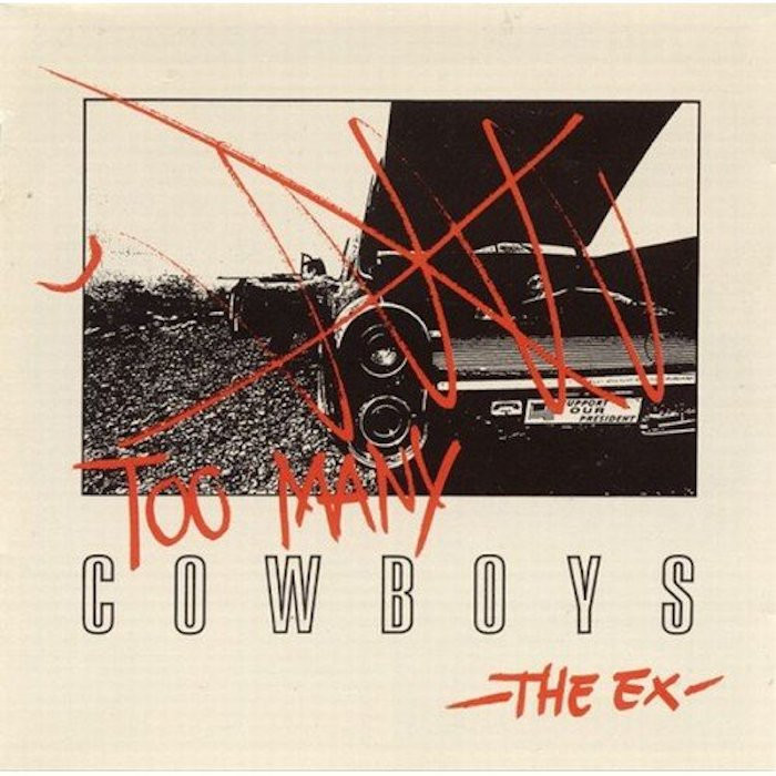 A white cover with a black and white photo of a car with its trunk open. The left door is open and a crouching man, in a shooting position with his eye in the sights, is using it to rest the barrel of his rifle. The photo is crossed out by hand with red lines, and the handwritten text "too many" is written above "cowboys".