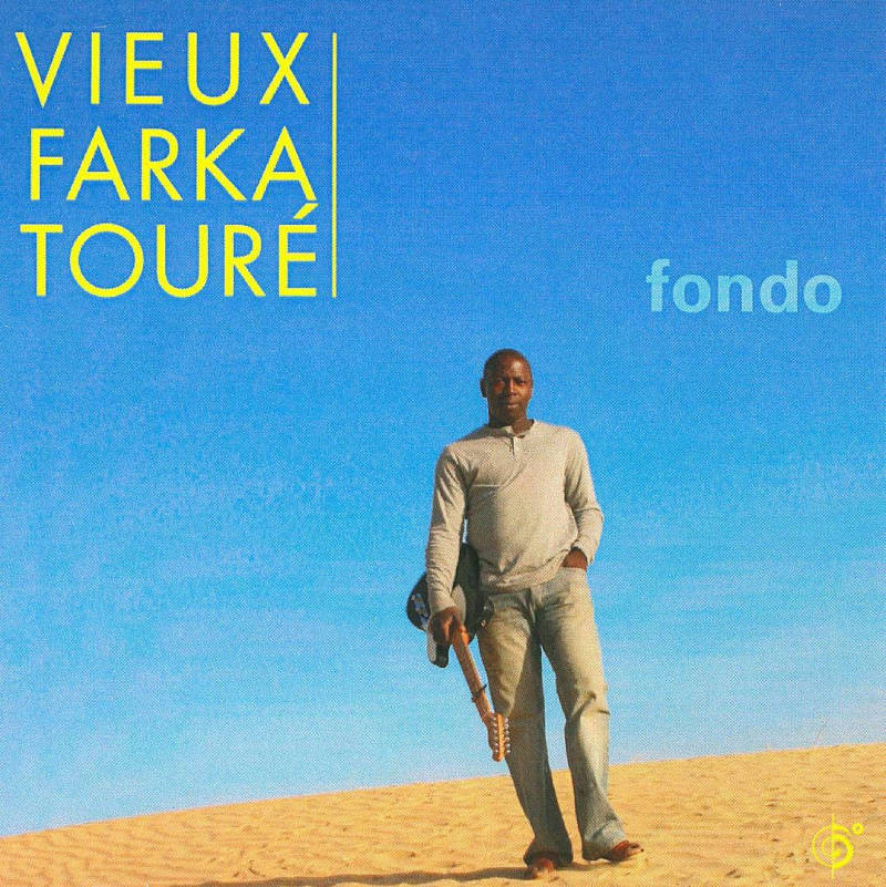Photo of Vieux Farka Touré walking on the sand in a long-sleeved T-shirt and jeans, an electric guitar in his right hand, in front of a beautiful azure blue sky.