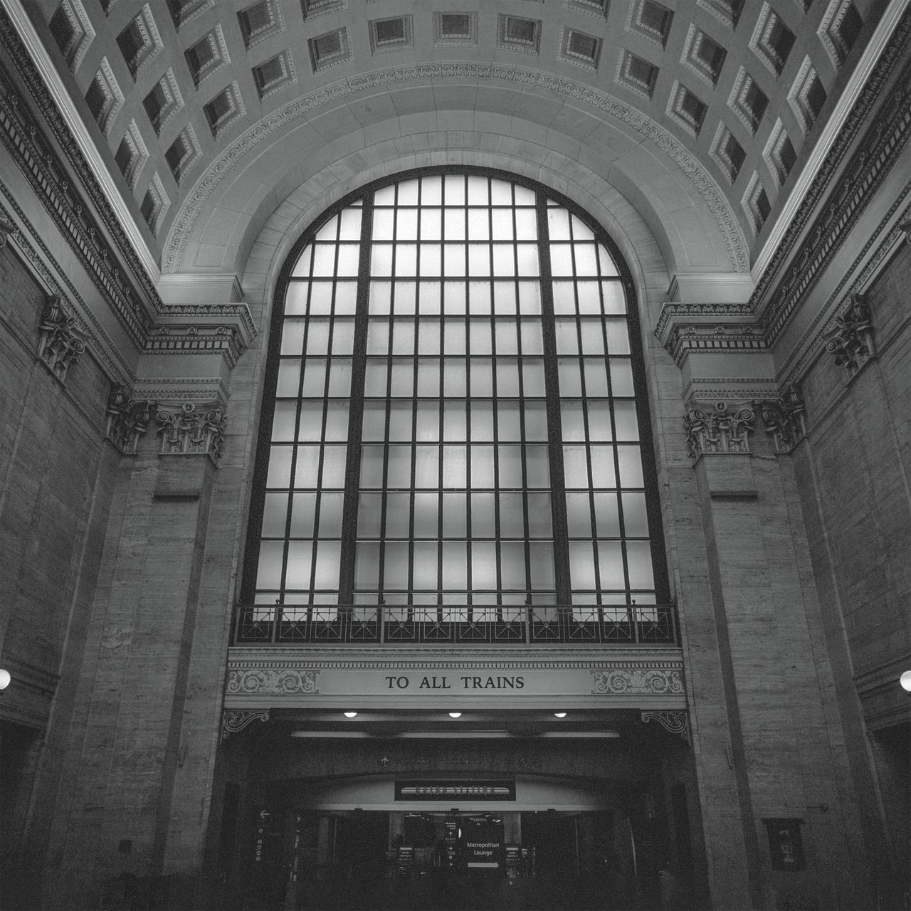Photo en noir et blanc en contre-plongée de l'intérieur d'une gare ancienne faite en pierres : le plafond est arrondi, une grande verrière descend de la voûte jusqu’à un passage pour les usagers, au-dessus duquel est indiqué : To all trains.