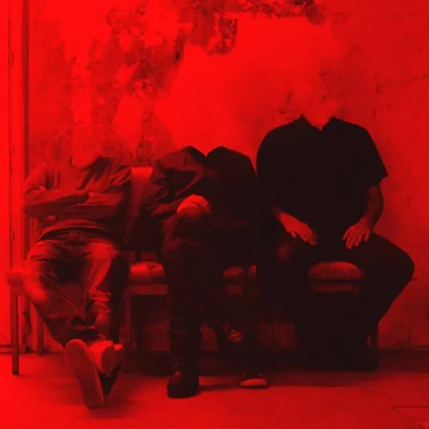 Photo noir et blanc, mais le blanc est remplacé par du rouge. 3 personnes sont assises avec 3 positions différentes sur une banquette le long d'un mur, les têtes sont effacées.