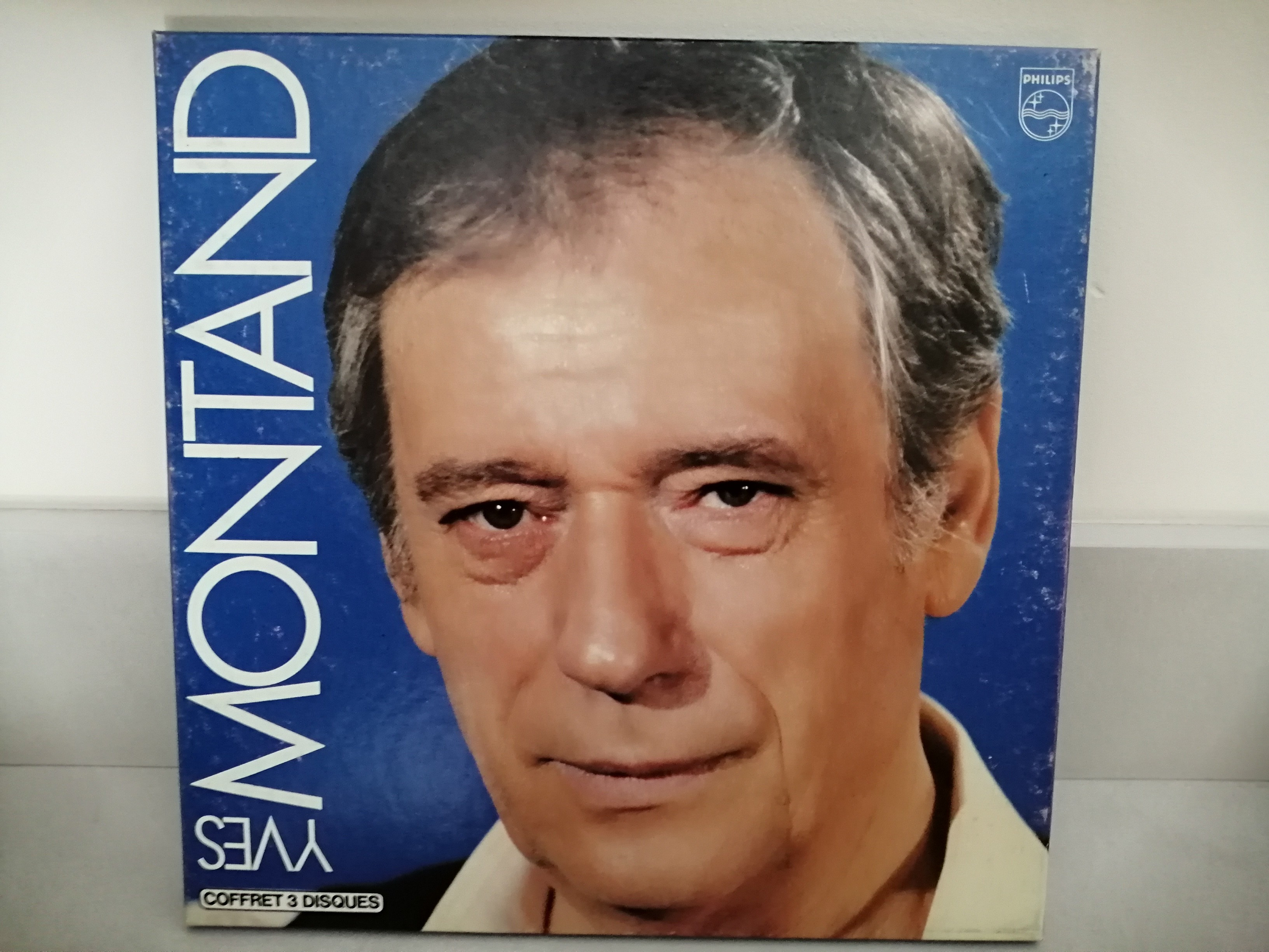 Sur un fond bleu roi le visage en couleur d'Yves Montand regardant fixement l'objectif. Sur le côté gauche en grandes lettres blanches "Yves Montand"