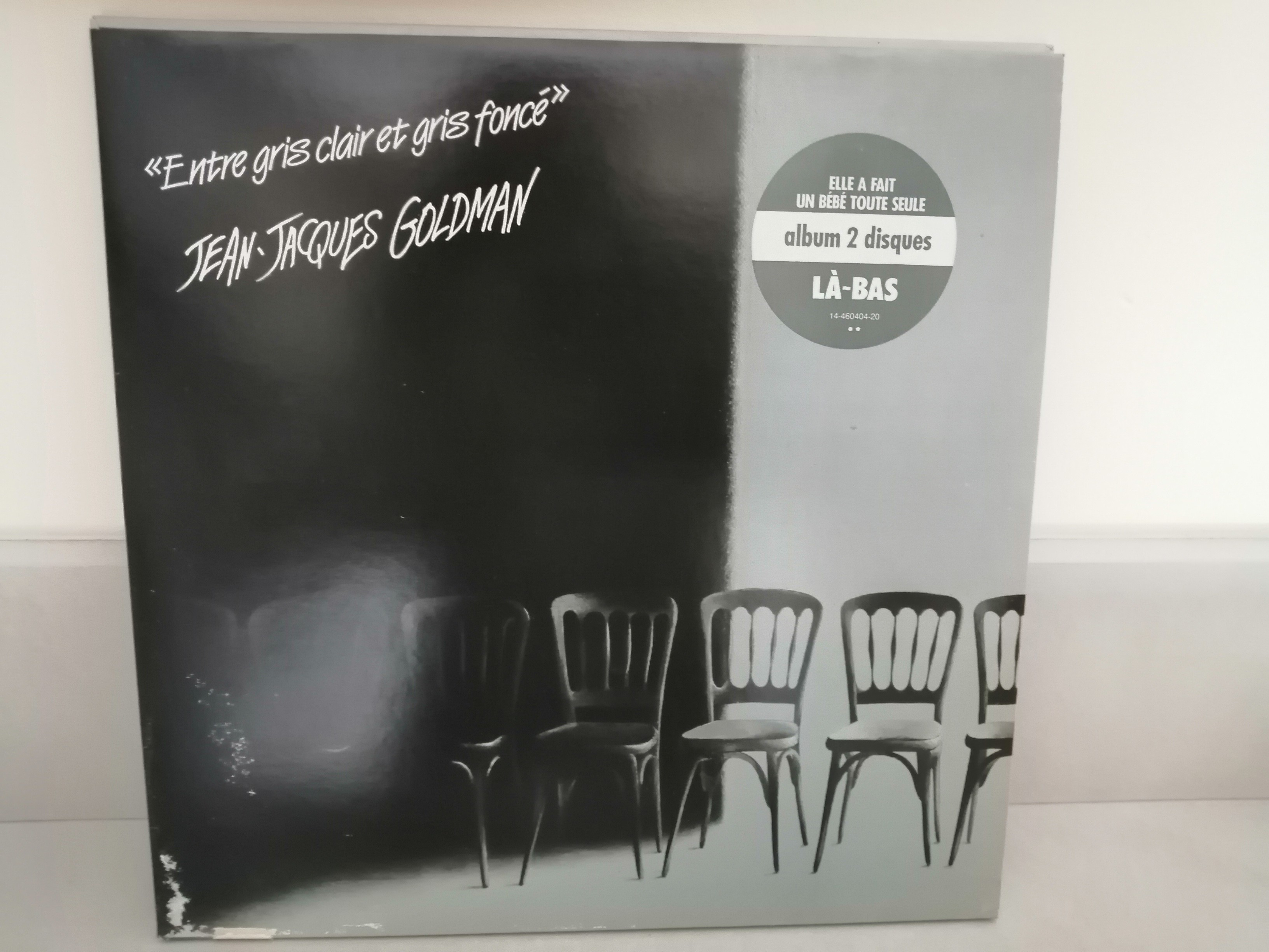 Sur un fond aux deux tiers noir sur la gauche et gris clair sur la droite, une série de 6 chaises en bois. En haut à gauche et en blanc l'inscription "Entre gris clair et gris foncé" Jean-Jacques Goldman.
