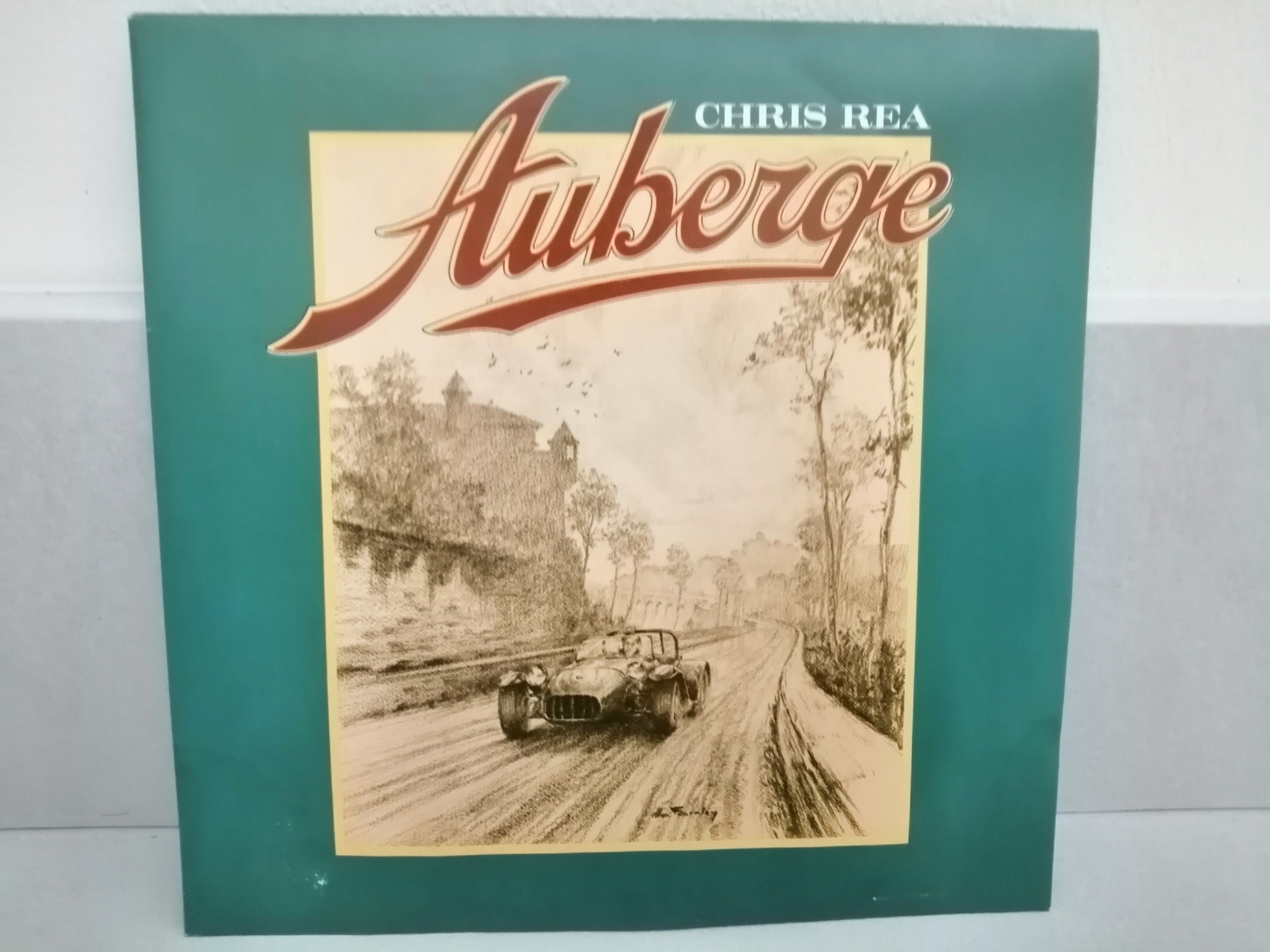 Un dessin monochone représentant une automobile (Lotus Seven) de 3/4 face sur une route. Au dessus le mot "Auberge" en rouge. Le tout est encadré de vert.