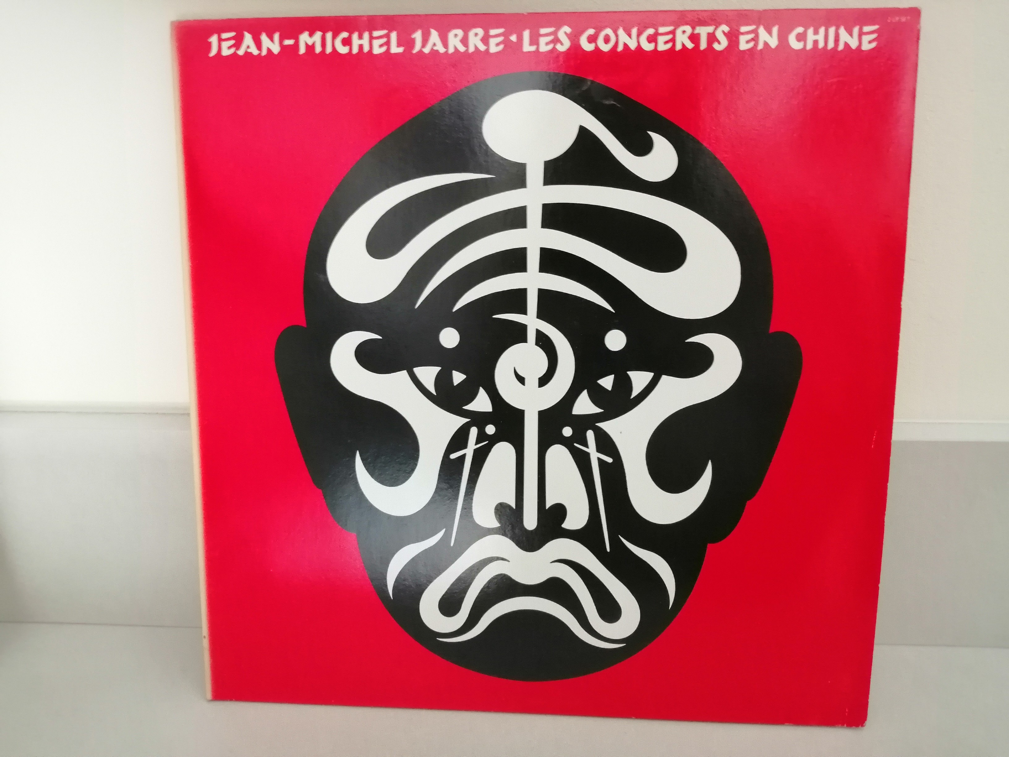 Sur un fond rouge vif, le dessin d'un visage noir avec des marques blanches rappelant les masques de théâtre chinois. En haut et en blanc l'inscription "Jean-Michel Jarre - Les concerts en Chine".