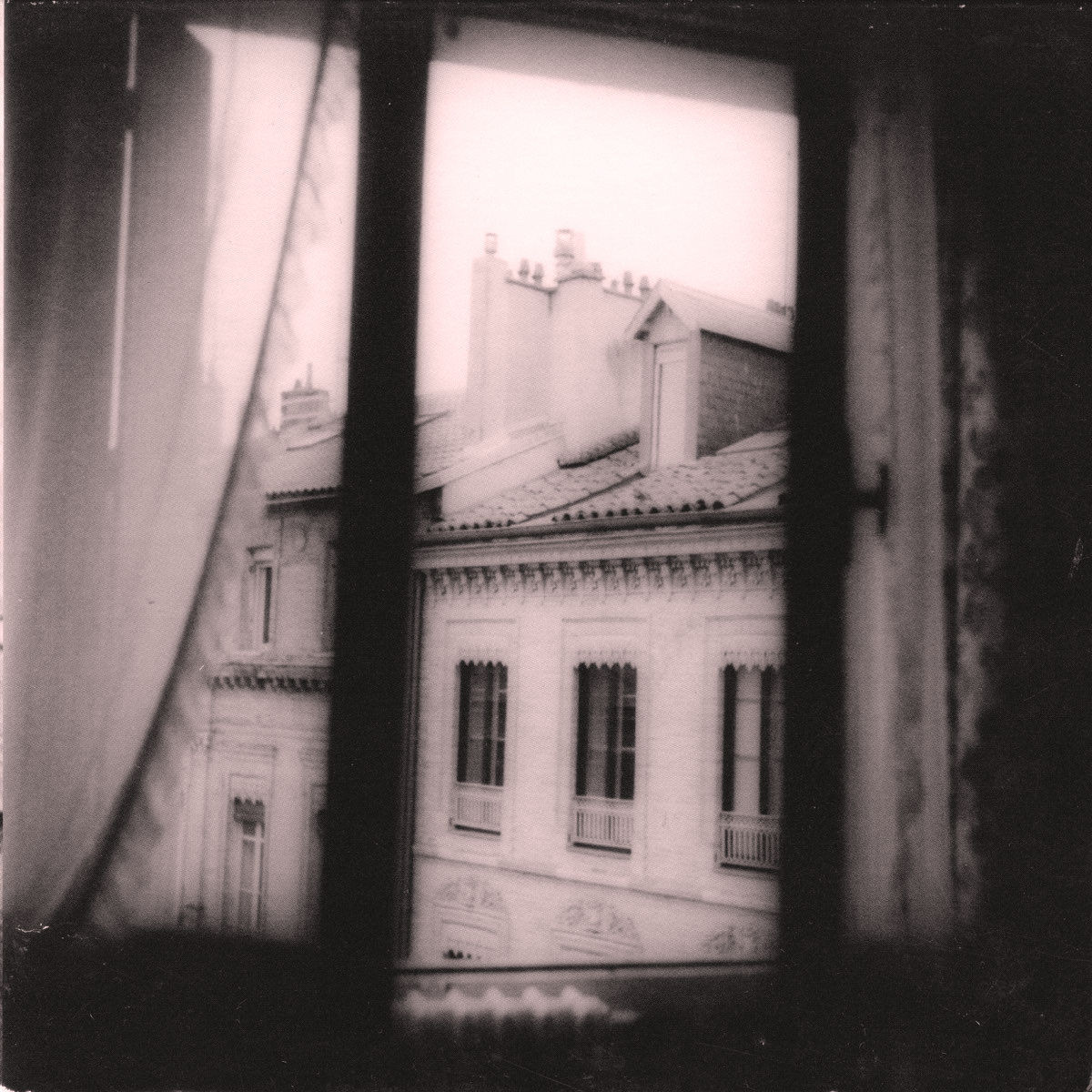 Photo en noir et blanc / sépia. Au travers d'une fenêtre entrouverte (le côté gauche est fermé, avec un rideau qui s'écarte en bas), on voit le haut de petits immeubles dans une rue : les fenêtres du dernier étage, les toits, cheminées. On aperçoit un radiateur sous la fenêtre