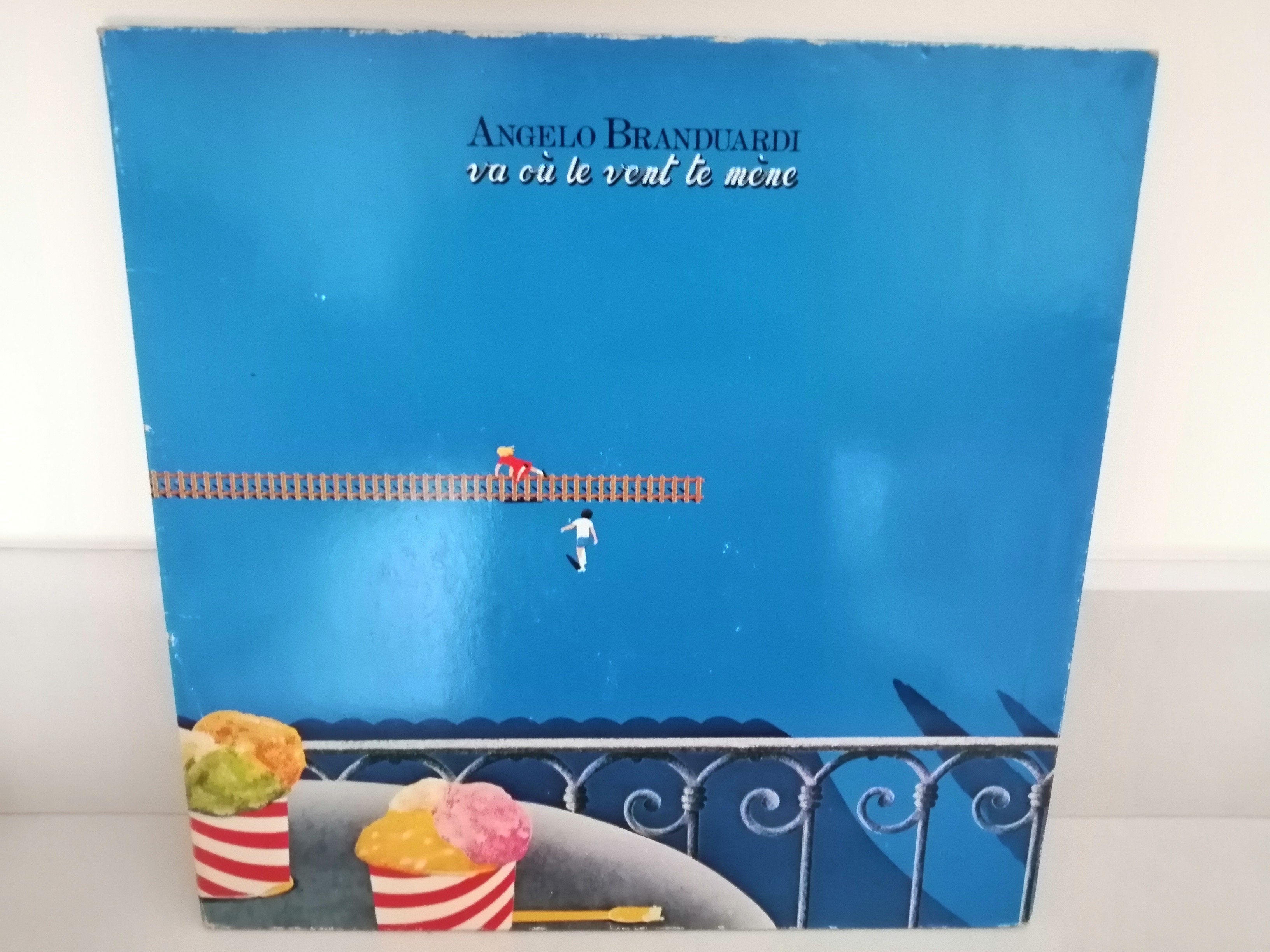 Une pochette de disque dessinée avec une vue depuis un balcon. On voit la rambarde en fer forgé en bas avec une table ronde sur laquelle sont posée deux pots contenant des boules de crème glacée. En contrebas du balcon un espace entièrement bleu avec un personnage en rouge qui enjambe une barrière. Un personnage en blanc le rejoint.
