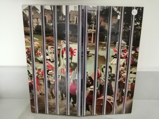 Une photo couleur extraite de l'épisode "Liberté pour tous" avec les Villageois, une fanfare faisant la fête autour de la place centrale du Village. Certains brandissent une pancarte "Vote n°2" et d'autres "Vote n°6". En superposition les barreaux d'une prison.
