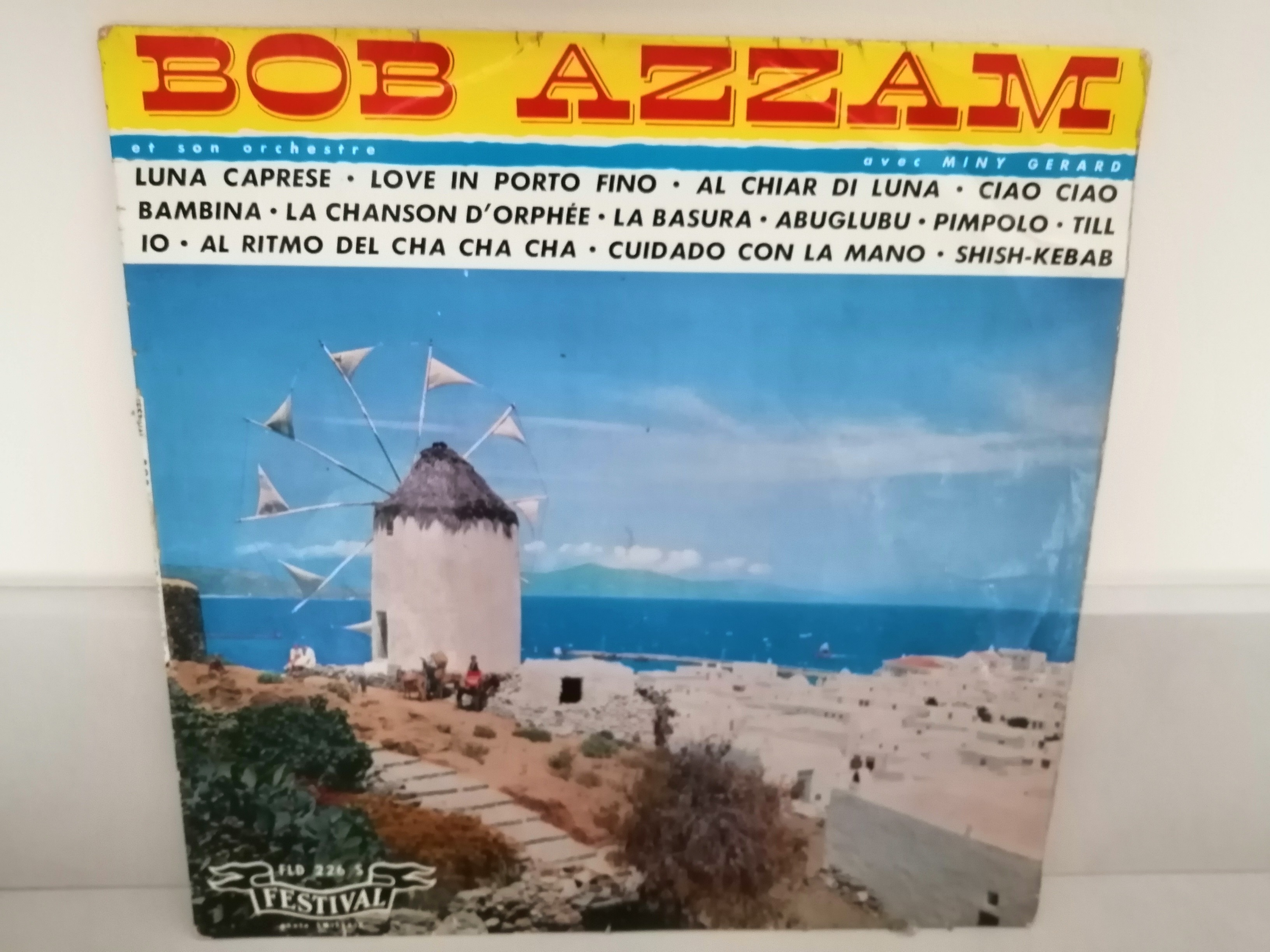 Un moulin à vent avec en arrière plan la mer et un grand ciel bleu avec quelques petits nuages. En haut est écrit "Bob Azzam" en grandes lettres rouges sur fond jaune. En dessous les titres des morceaux présents sur l'album en noir sur fond blanc.