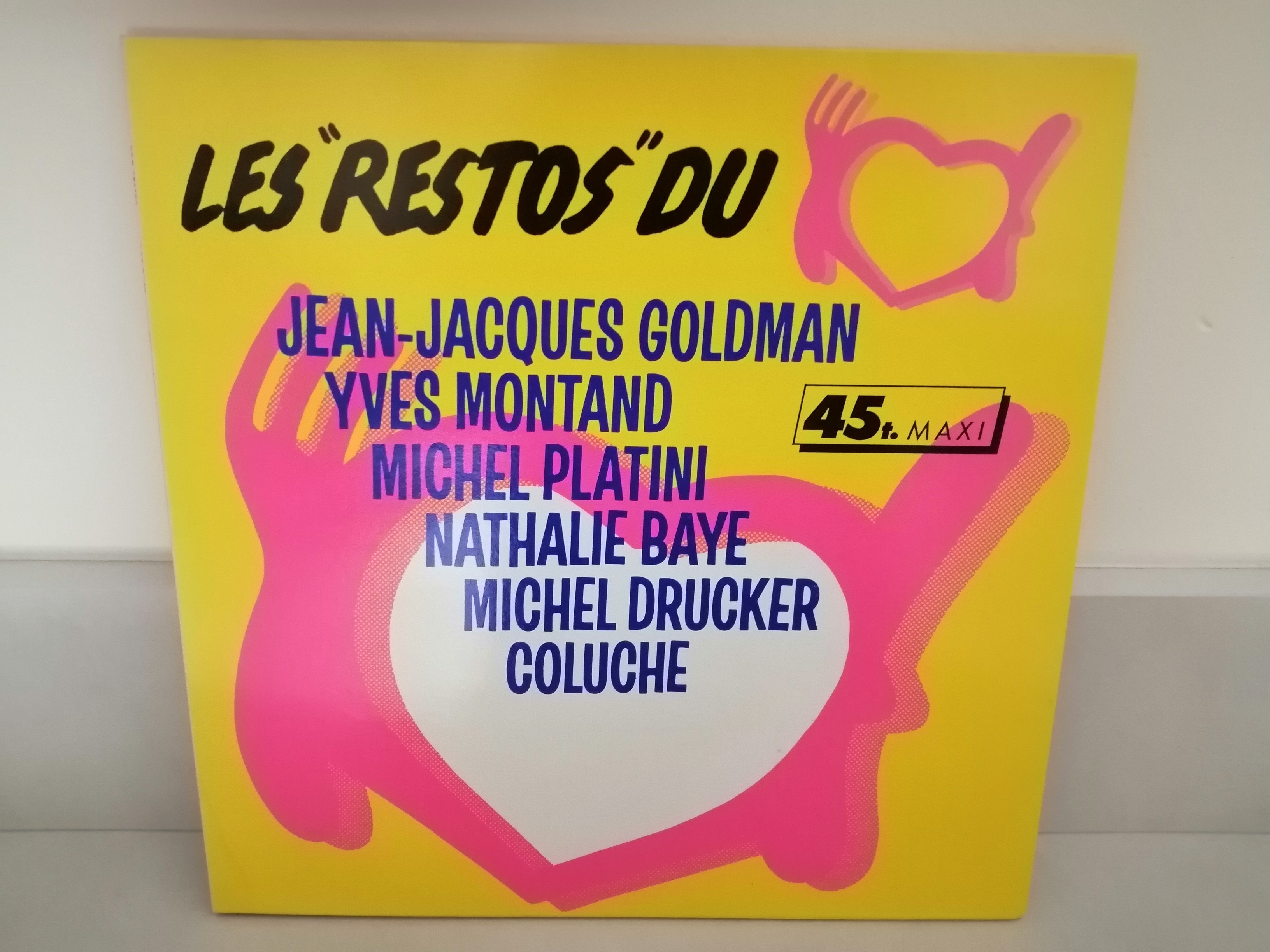 Sur un fond jaune fluo, un coeur rose vif avec de part et d'autre une fourchette à gauche et un couteau à droite de la même couleur.En haut le titre Les restos du Coeur et en dessous le nom des artistes ayant participé.