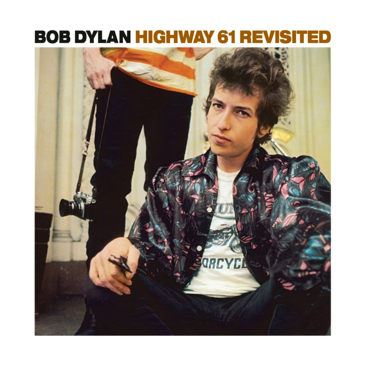 Une photo entourée d'un cadre blanc. Au premier plan de la photo Bob Dylan est assis et regarde l'appareil. Derrière lui on vois les jambes et le bas du torse de quelqu'un qui tiens un appareil photo a la main.Au dessus de la photo, le nom de l'artiste en noir et le tire de l'album en rouge