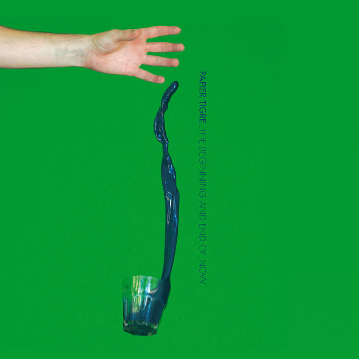 Against an all-green background, a hand opens, dropping a filled glass that leaves a column of dark blue liquid..