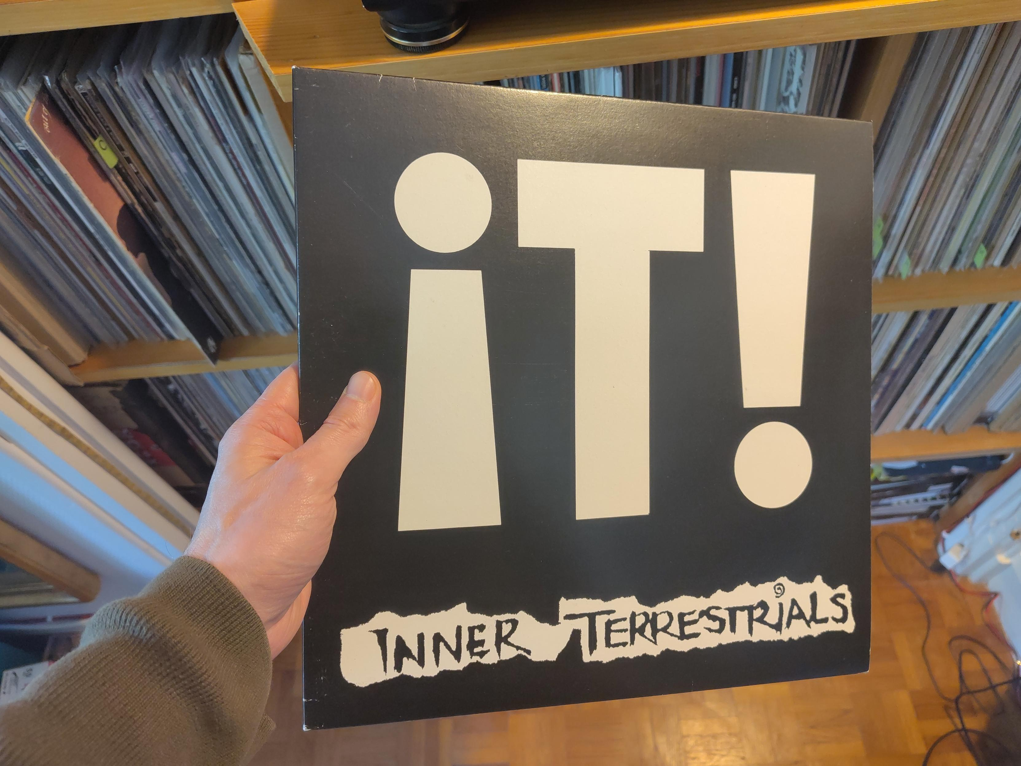 Le titre de l'album, IT! Couvre en très grosse lettres blanches la couverture qui pour le reste est noir. La composition joue avec le fait que la lettre « i » et le point d'exclamation ont exactement la même forme, simplement retournée sur la verticale. Le nom du groupe est lisible en bas, en noir sur un fond blanc qui imite une bande de papier découpé.