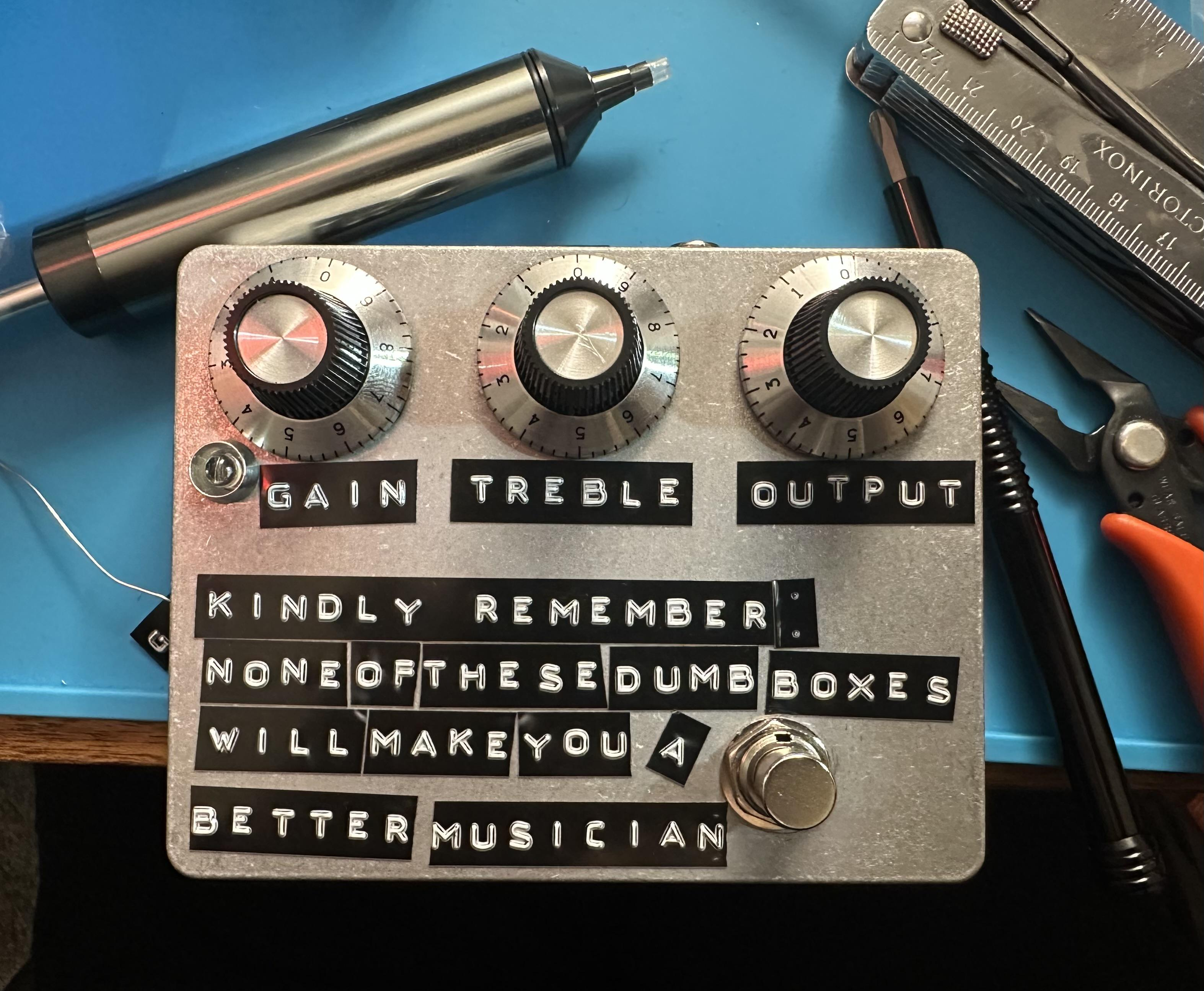un pédale d'effets avec une étiquette disant "Kindly remember that none of these dumb boxes will make you a better musician"