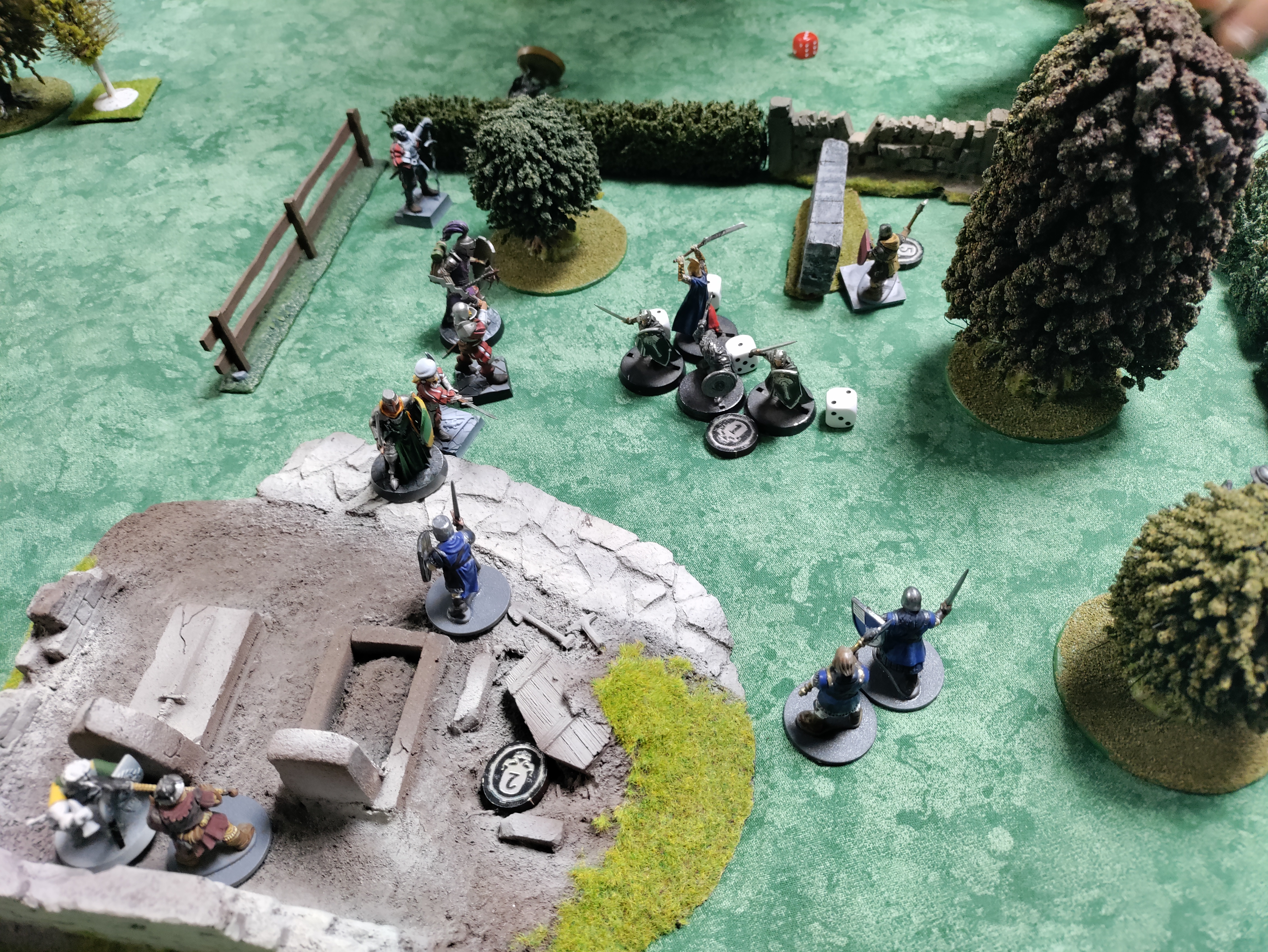 Une partie de jeu de figurine vue du dessus : sur une nappe verte, on voit un espace délimité par des barrières. Cet espace contiens des arbres, un petit cimetière et des murets, et on y voie évoluer des figurines de chevaliers en train de se battre, ainsi que quelques dés et marqueurs