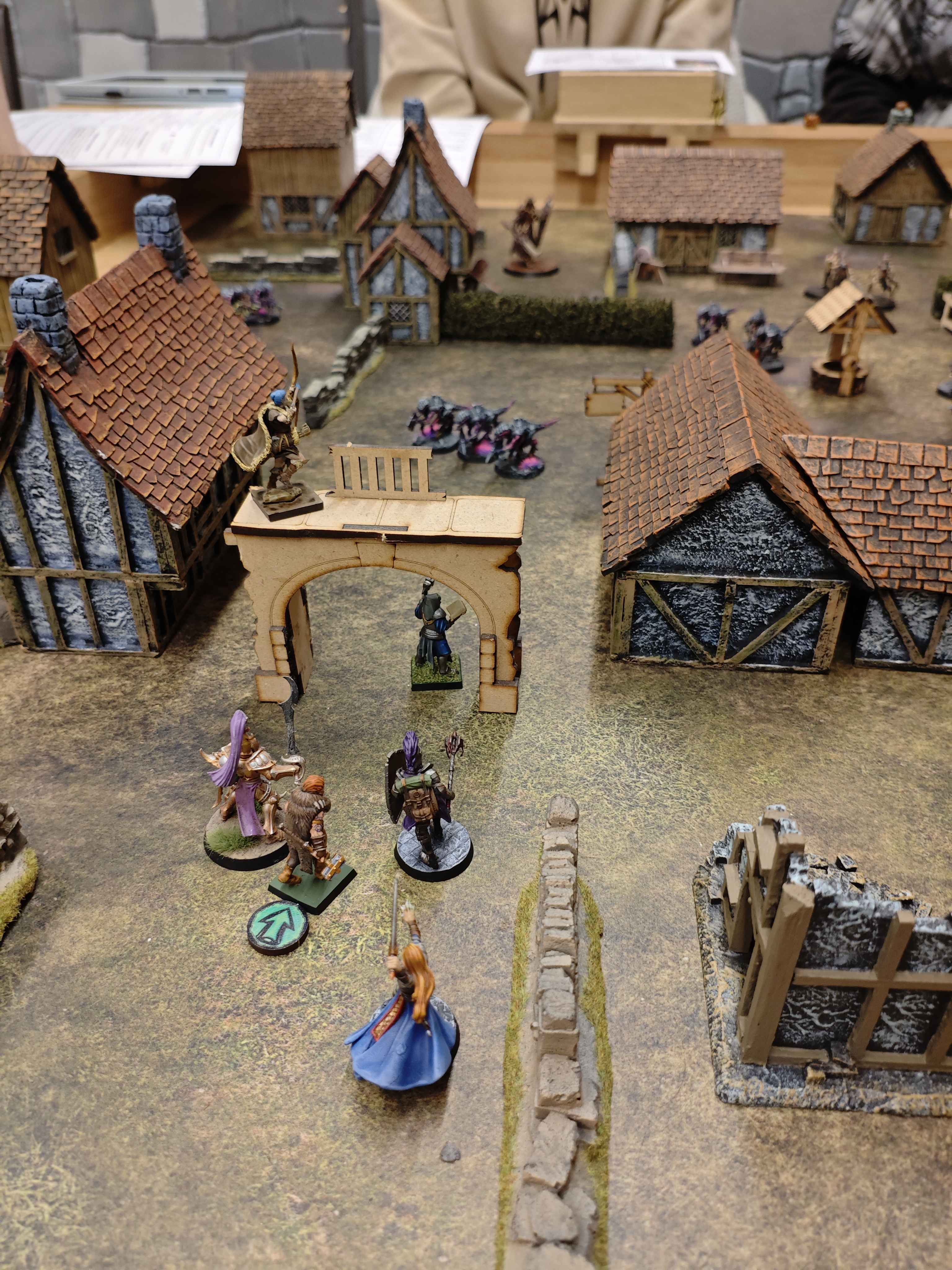 Une partie de jeu de figurines : on voit de dos 6 personnages qui s'apprètent à rentrer dans un village ou on distingue des monstres