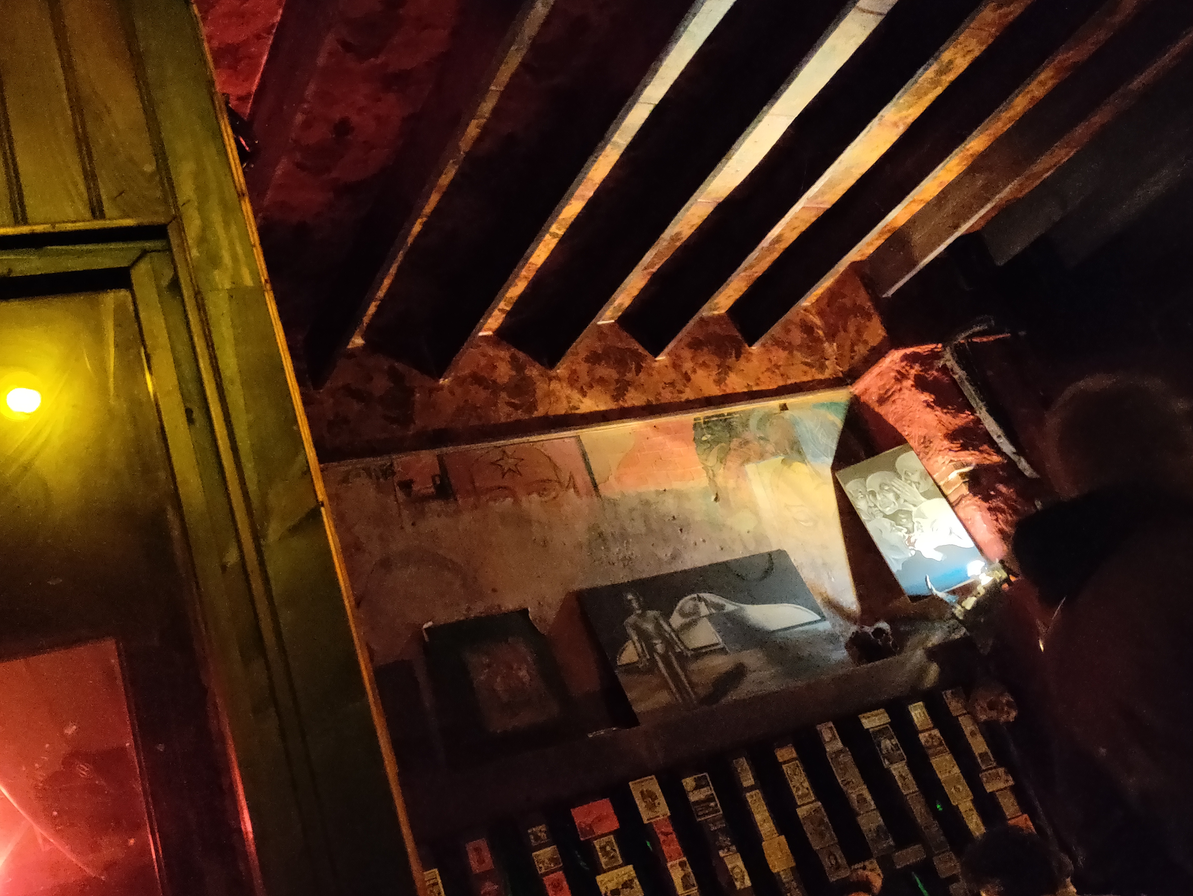 Photographie prise de travers de la salle d'un bar, peu éclairée, décrépite, avec des fresques effacées, des peintures psychédéliques et des dizaines de flyer sur les murs